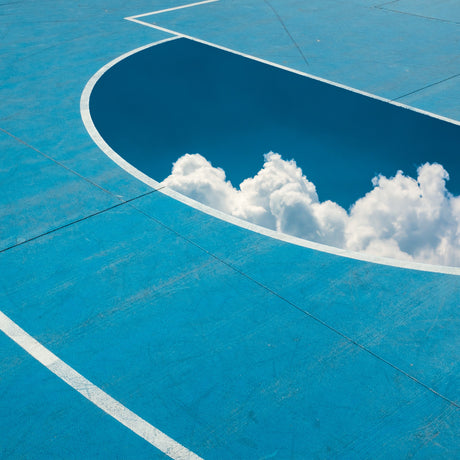 FREE THROW: This image shows a close-up of a blue basketball court with white lines. The court has a half-circle opening that reveals a bright blue sky with fluffy white clouds. The image is a surreal and creative representation of the connection between the mundane and the extraordinary.