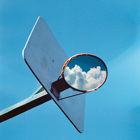 HOOP DREAMS: A basketball hoop is positioned against a bright blue sky, with the rim reflecting a puffy white cloud. The hoop is angled towards the viewer, with the backboard partially visible. The image captures a simple yet interesting perspective, highlighting the interplay between the man-made object and natural elements.