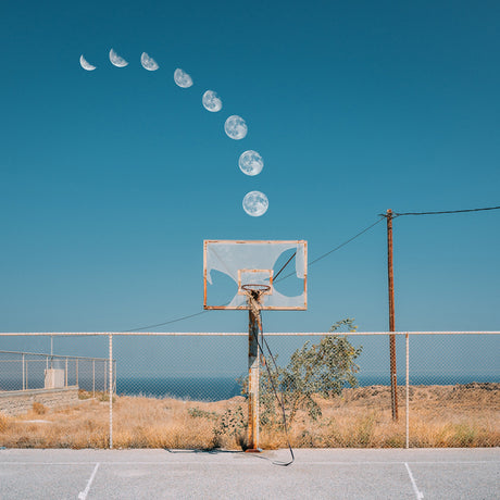 SWISH: A basketball hoop stands in a rural landscape with a chain link fence behind it. The hoop is old and rusty, and the net is missing. The moon is seen in the sky, and it appears to be sinking into the hoop. The scene is surreal and humorous, and it is a great example of how the ordinary can be made extraordinary with a little bit of creativity.