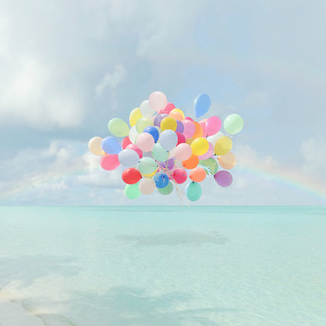 CATCH AND RELEASE: A cluster of colorful balloons floats high above a turquoise ocean with a bright blue sky and puffy white clouds in the background. The balloons are a mix of pinks, blues, yellows, and greens, creating a vibrant and cheerful scene.