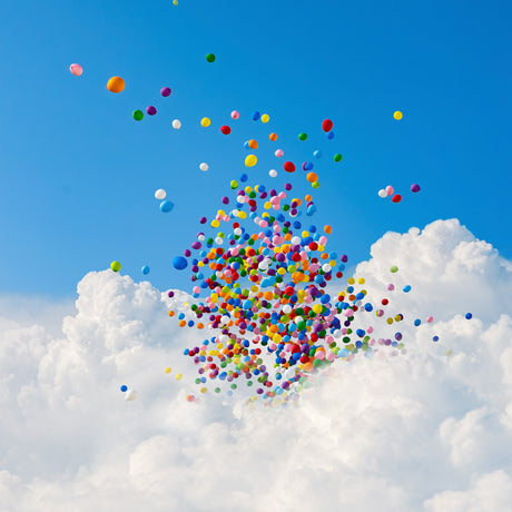 SUNBURST: This image shows a vibrant cluster of colorful balloons floating upwards in a bright blue summer sky. The balloons are surrounded by puffy, white clouds, creating a cheerful and celebratory scene.