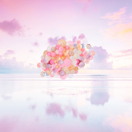 SUNSET RISING: A large cluster of pastel pink, yellow, and white balloons floats gently over a calm, sandy beach at sunset. The sky is filled with soft, pink and purple clouds, creating a dreamy and romantic atmosphere. The reflection of the balloons and clouds is visible in the water, adding to the enchanting beauty of the scene.