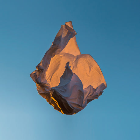 VIENTO 02: A white plastic bag floats in the air against a blue sky, illuminated by the warm light of the setting sun. The bag is crumpled and distorted, creating an abstract shape against the background.