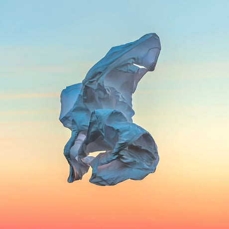 VIENTO 04: A blue plastic bag floats in the air against a vibrant sunset sky. The bag is caught in the wind and billows out, creating a surreal and abstract shape. The colors of the sky are a combination of orange, pink, and blue, adding to the beauty of the image.