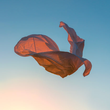 VIENTO 05: A white fabric billows and dances in the wind against a clear blue sky with an orange sunset in the background. The fabric is caught by the wind, creating a dynamic and graceful shape.