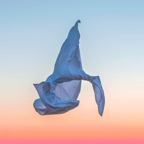 VIENTO 08: This image features a blue fabric piece flowing in the wind against a beautiful sunset sky. The fabric is caught in a dynamic motion, creating a sense of movement and freedom. The colors of the sunset, with hues of pink, orange, and blue, create a striking contrast against the fabric, making it a visually interesting composition.