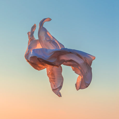 VIENTO 09: A white piece of fabric, possibly silk, flows gracefully in the wind against a backdrop of a colorful sunset sky. The fabrics movement creates a sense of freedom and lightness.