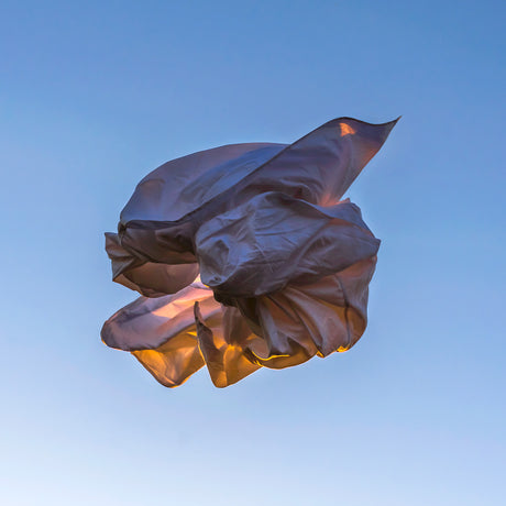 VIENTO 15: A close-up shot of a piece of white fabric billowing in the wind, backlit by the setting sun. The fabric is caught in a swirling motion, creating an abstract and dynamic form. The sky is a clear blue, providing a serene backdrop for the vibrant and energetic fabric.