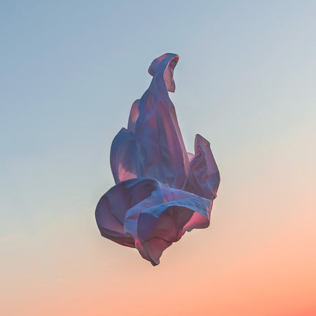 VIENTO 18: A piece of purple fabric floats in the air against a backdrop of a vibrant pastel sky. The fabric is caught in the wind, creating a dynamic and abstract shape, and the light from the setting sun casts a warm glow on the scene.