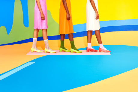 BIG SUN: Four women in colorful dresses and socks stand side-by-side on a platform. The platform appears to float in front of a colorful wall. The women are all looking straight ahead with serious expressions. They are wearing pink, orange, and white dresses, and their socks are yellow, green, and white. The background is a bright, abstract wall with shades of green, yellow, and blue.