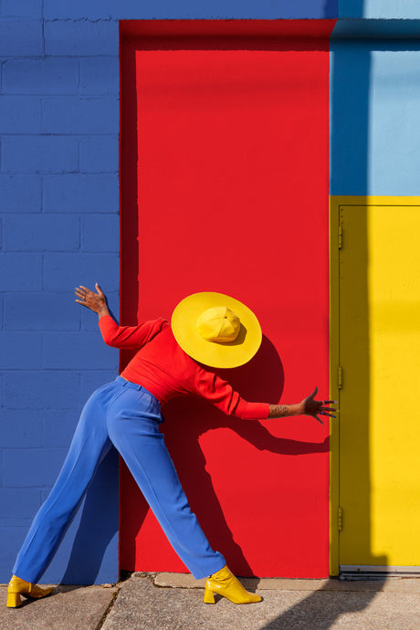 PRIMARY POSE: This image shows a woman wearing a red shirt, blue pants, yellow hat, and yellow boots leaning against a red door. She is standing in front of a blue wall with a yellow door to her right. The womans pose is dynamic and energetic, creating a sense of movement and energy.