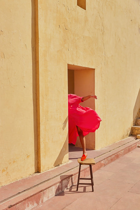 SUNDIAL: A woman in a vibrant pink dress stands on a wooden stool outside a yellow building. The dress billows in the wind, and her red shoes are visible on the stool. The setting appears to be a traditional Indian location.