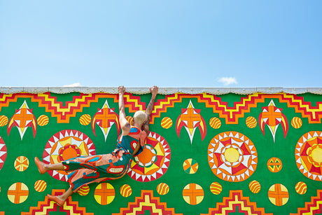 THE SWING: This image shows a woman hanging upside down from a colorful wall mural, with her legs dangling over the edge. The mural features a bright green background with geometric patterns in red, orange, yellow, and white. The woman is wearing a green, orange, and white patterned jumpsuit, and she appears to be having fun.