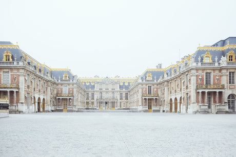 VERSAILLES: A wide shot of the Palace of Versailles in France, taken on a cloudy day. The iconic building is seen from the courtyard, with a few people walking in the foreground.