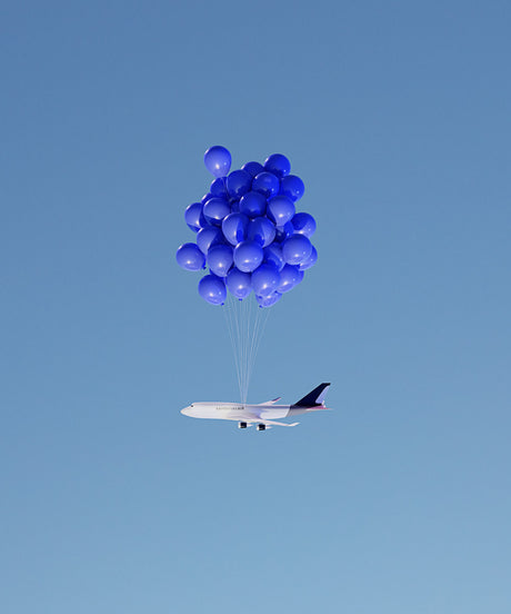 BALLOON FLIGHT 1: A white airplane with blue and black markings is suspended in mid-air by a cluster of blue balloons against a bright blue sky. The airplane appears to be a commercial airliner, and the balloons are a vibrant blue color, adding a whimsical touch to the scene.