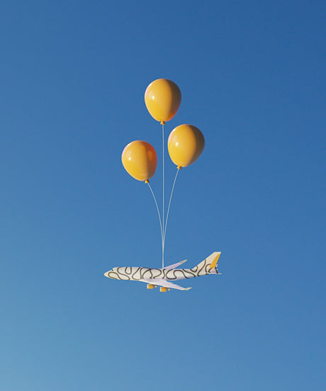 BALLOON FLIGHT 4: A white airplane with black abstract patterns on its exterior is being held aloft by three yellow balloons against a clear blue sky. The airplane appears to be a toy or a model, and the balloons are a vibrant yellow. The image captures a whimsical and playful moment of flight.