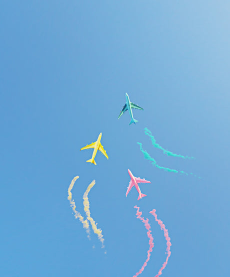 PASTEL FLIGHT 1: Four airplanes fly in formation, leaving colorful smoke trails behind them as they perform aerial stunts against a clear blue sky. The airplanes are yellow, blue, and pink.