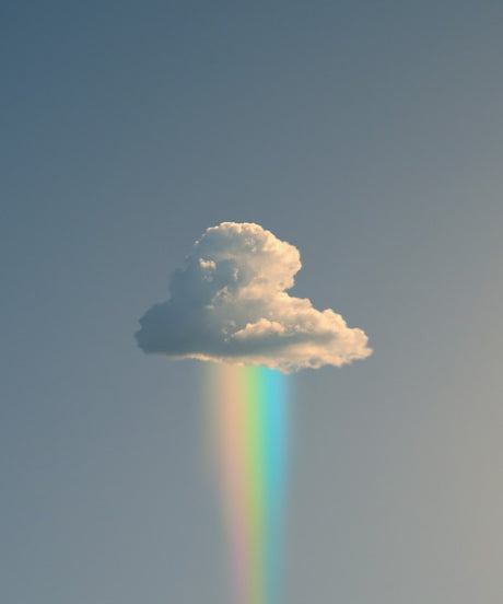 RAINBOW RAIN: A single, fluffy white cloud hangs in a vast blue sky. A thin, pastel rainbow extends downwards from the cloud, illuminating the sky below. The image captures a serene and beautiful moment in nature.