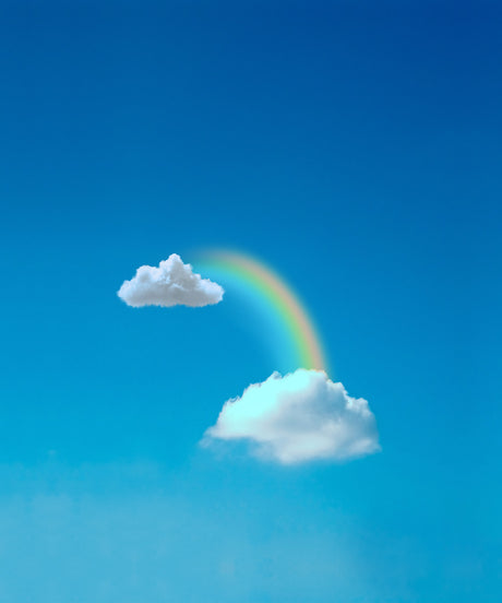 RAINBOW STEPS: A single rainbow arches between two white clouds in a bright blue sky. The rainbow is visible against the clear blue sky, with the white clouds framing it on either side. The image captures a moment of serenity and beauty in nature.