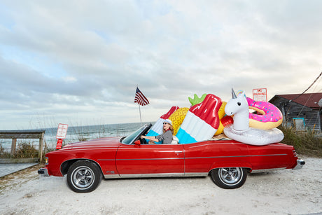 FAST AND FLOATY: A woman drives a classic red convertible car filled with inflatable floats, including a unicorn, a popsicle, and a donut, on a sunny day at the beach. There is an American flag in the background. The car is parked near a wooden walkway and the ocean is visible in the distance.