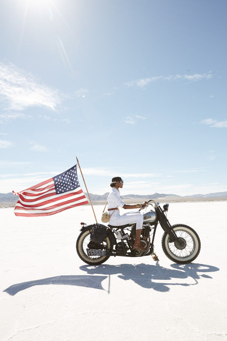 DESERT RIDE: A woman in white pants and a white shirt rides a motorcycle with an American flag attached to the back. The photo was taken on a sunny day at the Bonneville Salt Flats.