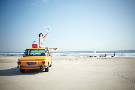 PINWHEEL GIRL 1: A woman is sitting on the roof of a yellow car, holding a pinwheel. She is wearing a white dress with red polka dots and red shoes. The car is parked on a sandy beach with the ocean in the background. There is a man walking with a dog in the distance. The sky is blue and the sun is shining.