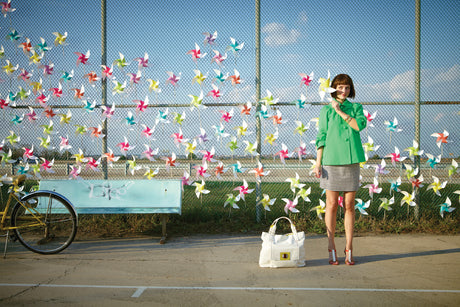 PINWHEEL GIRL 2: A woman in a green jacket and black and white skirt stands in front of a chain link fence covered in colorful pinwheels. She is holding one pinwheel in front of her face and smiling at the camera. There is a bench behind her and a bicycle to the left.