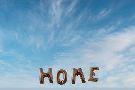 HOME: A photograph of a clear blue sky with white clouds. The word HOME is spelled out in large golden balloons floating in the sky.