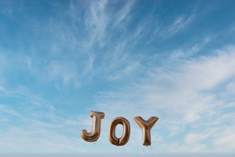 JOY: A photograph showcasing three golden balloon letters spelling out the word JOY against a backdrop of a clear blue sky with wispy white clouds. The balloons are slightly deflated and appear to be floating in the air.