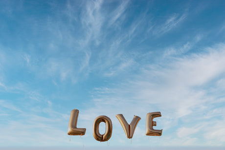 LOVE: A photograph of four large gold foil balloons spelling out the word LOVE against a backdrop of a clear blue sky with wispy white clouds. The balloons are floating in the air, with their strings trailing behind them.