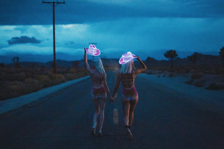 NEON DREAM II: This image shows two women, walking away from the camera on a desert road at dusk. They are wearing neon cowboy hats and similar outfits. The image has a moody and mysterious feel, with the women disappearing into the darkness of the desert.