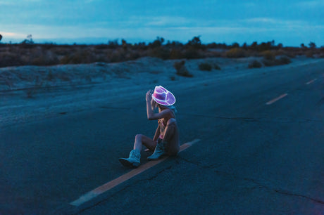 NEON DREAM: A woman wearing a neon pink cowboy hat sits on a deserted road in the middle of the desert. The sky is a pale blue, indicating dusk. The woman is facing away from the camera, but her hand is raised as if to signal a car.