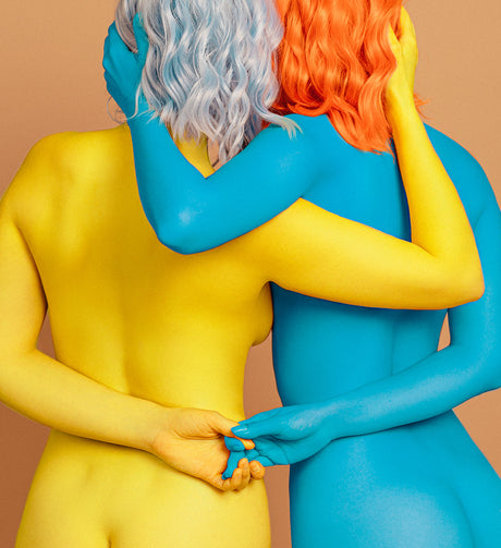 TOGETHER: This image shows two women standing close together, embracing. One woman is painted completely blue, and the other is completely yellow. They are both wearing wigs, one blue and one orange. The background is a light tan.