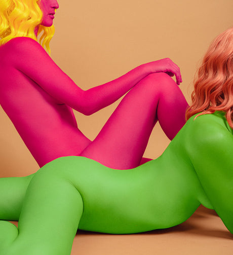 WAITING: This image features two women, one painted pink and one painted green, posing together in a studio setting. The women are both nude, but their bodies are painted in vibrant colors, creating a striking visual contrast. The woman in pink is sitting with her legs crossed, while the woman in green is lying on her side, facing the camera. Their poses are both relaxed and suggestive, creating a sense of intimacy and connection.