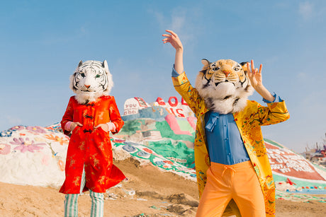 WILD THINGS II: This image shows two people wearing tiger masks, one in a red coat and the other in a yellow coat, posing in front of the Salvation Mountain art installation. They are both looking at the camera and smiling. The background is a bright blue sky and a desert landscape.