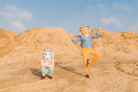 WILD THINGS I: This image shows two people wearing tiger masks posing in a desert landscape. One person stands on one leg with their arms outstretched, while the other person sits with their arms crossed. The desert landscape is characterized by sandy hills and a blue sky.