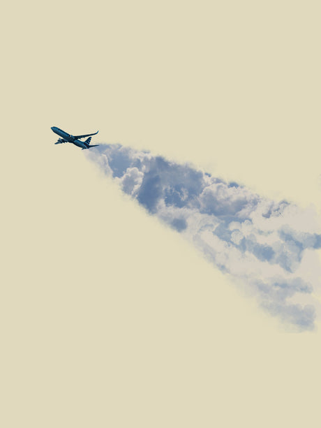 SOFT SKIES: A single airplane flies through a cloud formation, leaving a trail of condensation behind it. The sky is a pale blue with a light yellow hue.