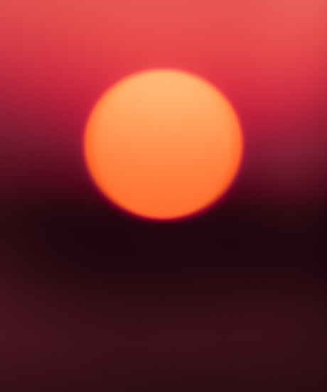 ENDLESS SUMMER NO8: A close-up, blurry photo of the setting sun, capturing its bright orange glow against a dark red sky. The focus is soft, emphasizing the warm, ethereal quality of the moment.